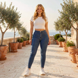 A woman stands on a sunlit stone terrace lined with potted olive trees, wearing a white cropped short-sleeve top, blue high-waisted skinny jeans, and white sneakers.