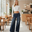 A woman stands in a bright cafe wearing dark blue wide-leg jeans, a white long-sleeve crop top, and white slip-on shoes.