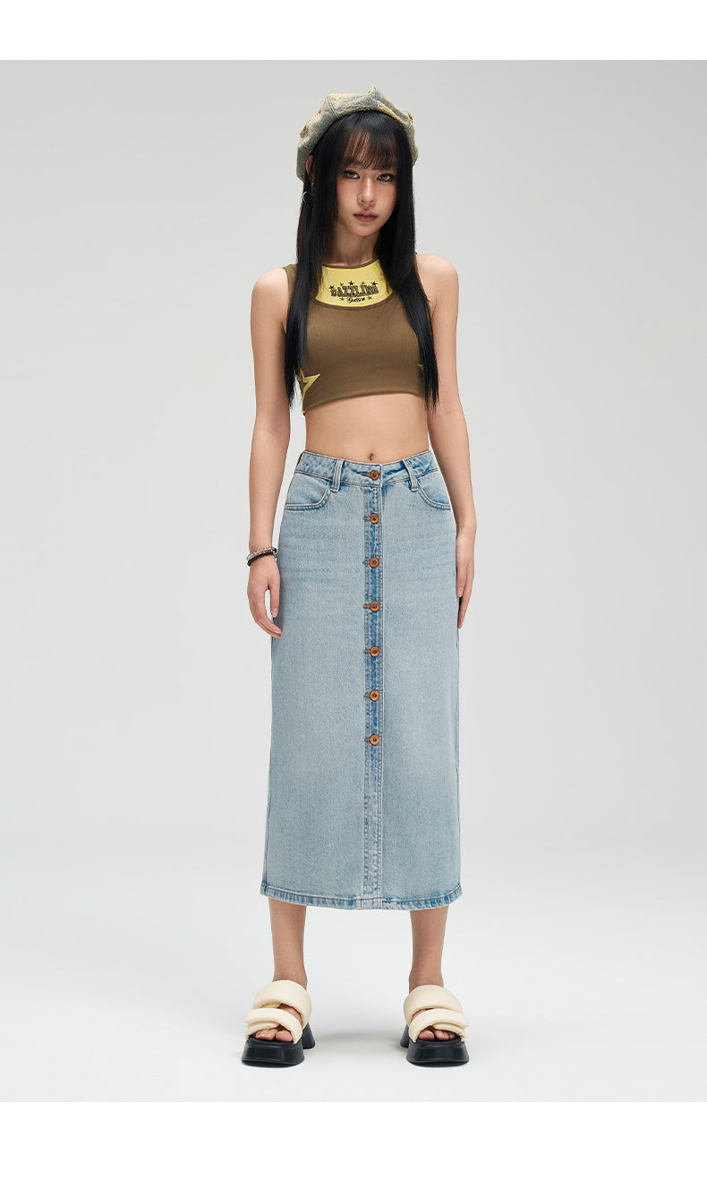 A model stands against a light gray studio background wearing a light-wash denim midi skirt with a button-front closure, a brown sleeveless crop top, and beige slide sandals.