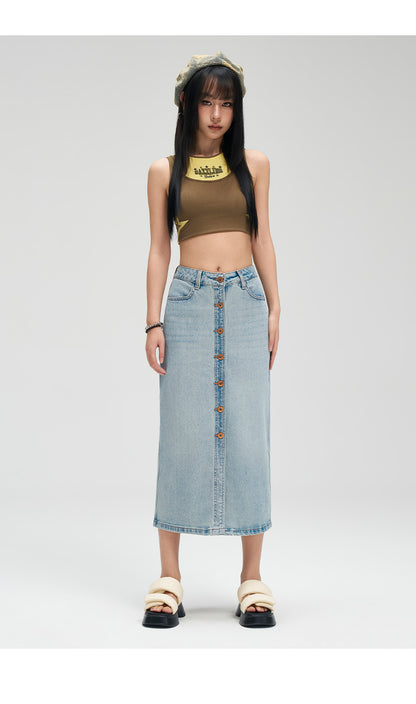 A model stands against a light gray studio background wearing a light-wash denim midi skirt with a button-front closure, a brown sleeveless crop top, and beige slide sandals.