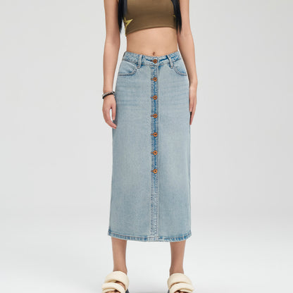 A model wearing a light-wash blue denim midi skirt with a button-down front, paired with a brown crop top and beige slide sandals, against a plain light gray studio background.