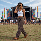 Woman at a music festival wearing sunglasses and a white tank top with brown jeans, with a stage and colorful flags in the background.