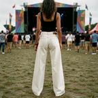 Person in white wide leg denim jeans wearing a black top and white pants at a music festival with colorful flags and stage in the background.