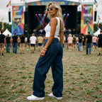 Woman at a music festival wearing sunglasses and a white top with blue wide leg low rise jeans, with a stage and crowd in the background.