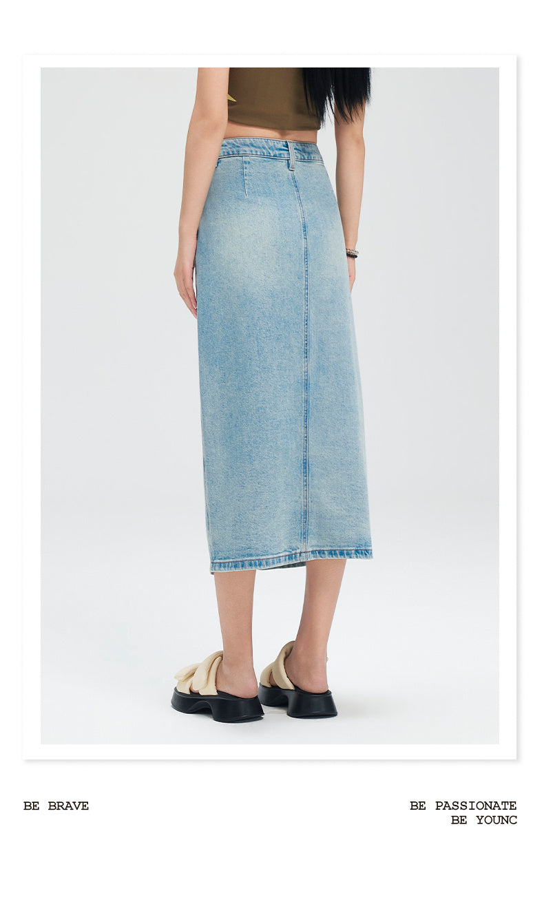 A model is shown from the waist down wearing a light blue denim midi skirt, paired with a dark olive top and beige-and-black platform mules, set against a plain light gray studio background.