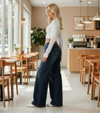 A woman stands in a bright, modern cafe wearing dark blue wide-leg jeans, a white long-sleeve crop top, and white sneakers.